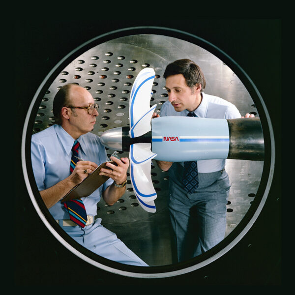 Two men standing next to a plane propeller in a room with metal walls.