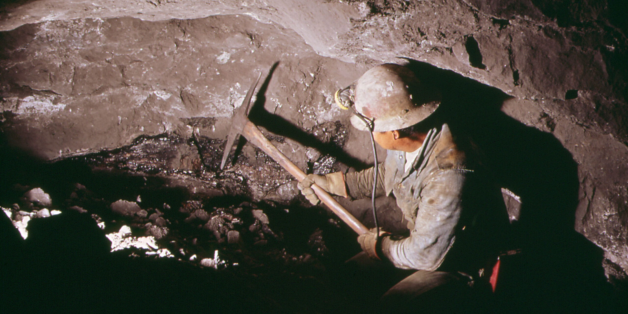 A man wearing a hardhat and headlamp wielding a pickaxe underground.