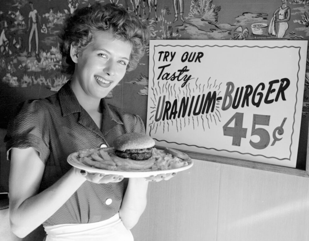 Woman holding a plate containing a hamburger and fries. A sign behind her reads: “Try our tasty uranium-burger.”