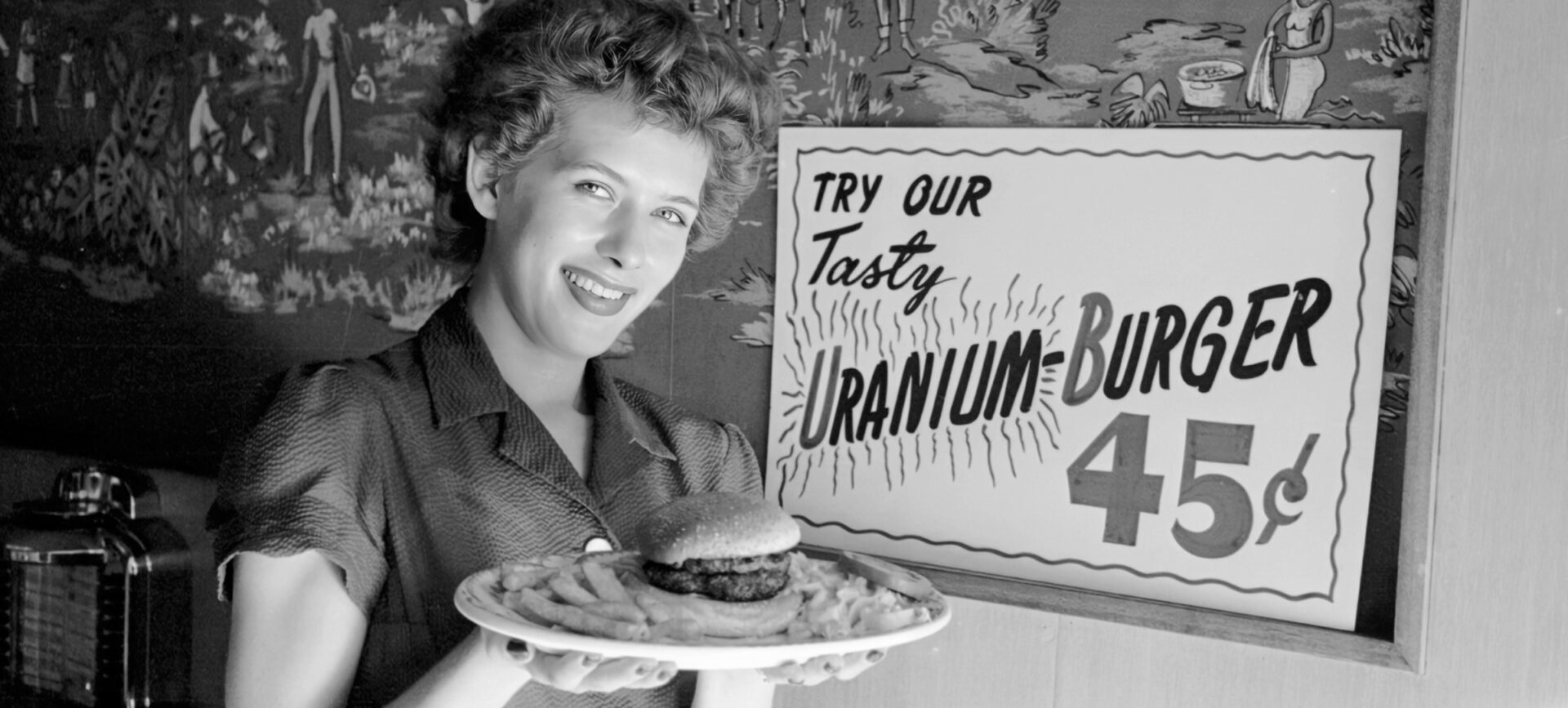 Woman holding a plate containing a hamburger and fries. A sign behind her reads: “Try our tasty uranium-burger.”