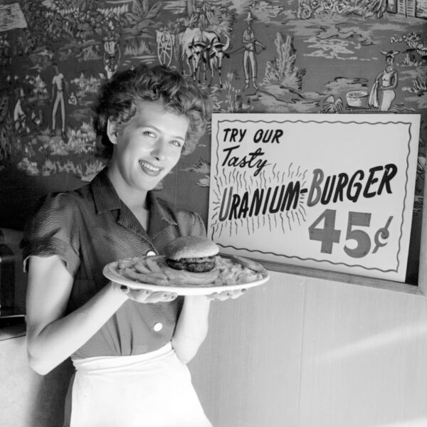 Woman holding a plate containing a hamburger and fries. A sign behind her reads: “Try our tasty uranium-burger.”