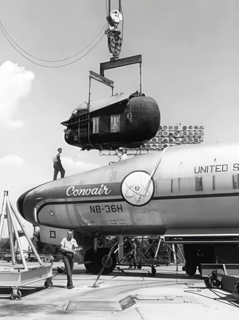 A large capsule being lowered into the nose of a jet aircraft. A stylized decal of an atom is visible on the side of the plane.