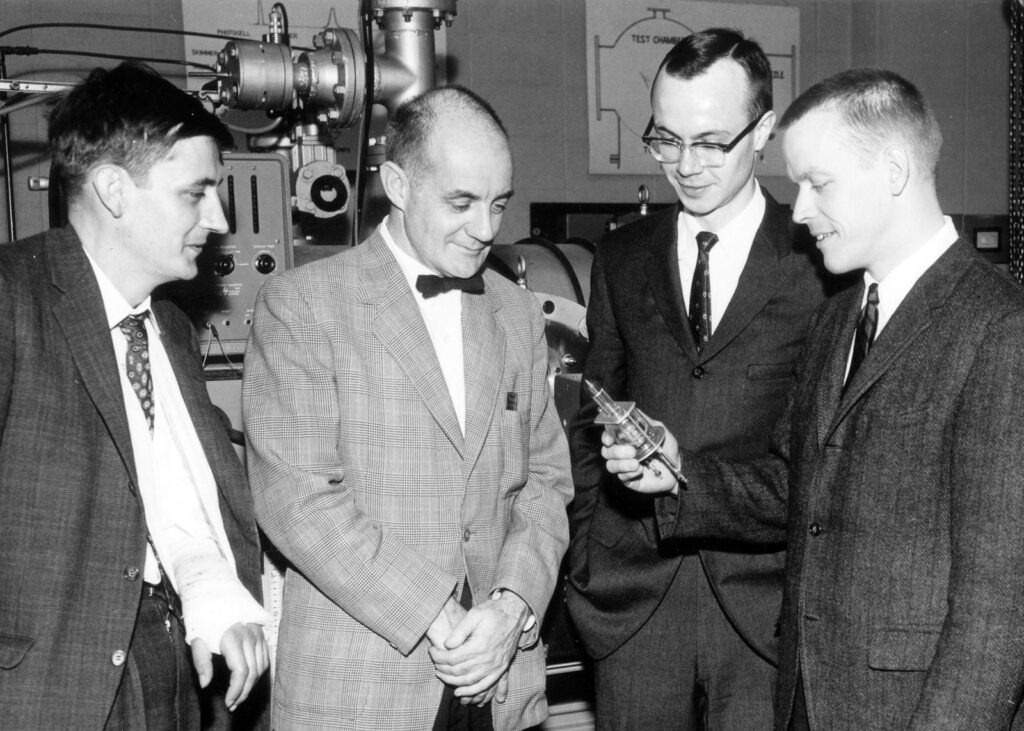 Four men in suits inspecting a small metal device in a laboratory, with a large machine in the background