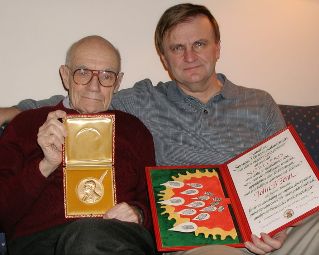 Two men sitting on a couch holding a medal and a diploma