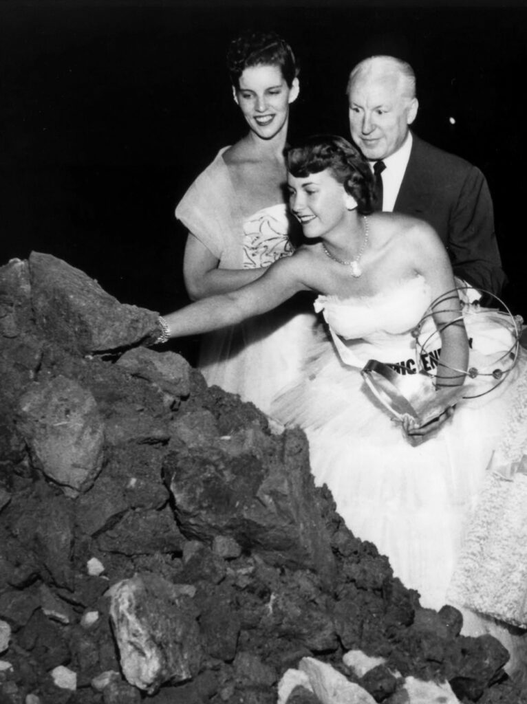 Woman in a white gown reaching out to touch a large pile of rocks. A man and women look on.