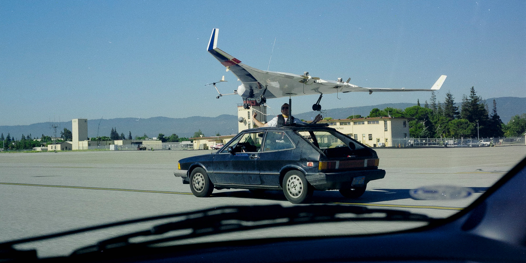 A plane attached to the roof of a vehicle on a runway. A man is standing up inside the car, with half of his body through the moonroof, and grasping to wires attached to the plane.