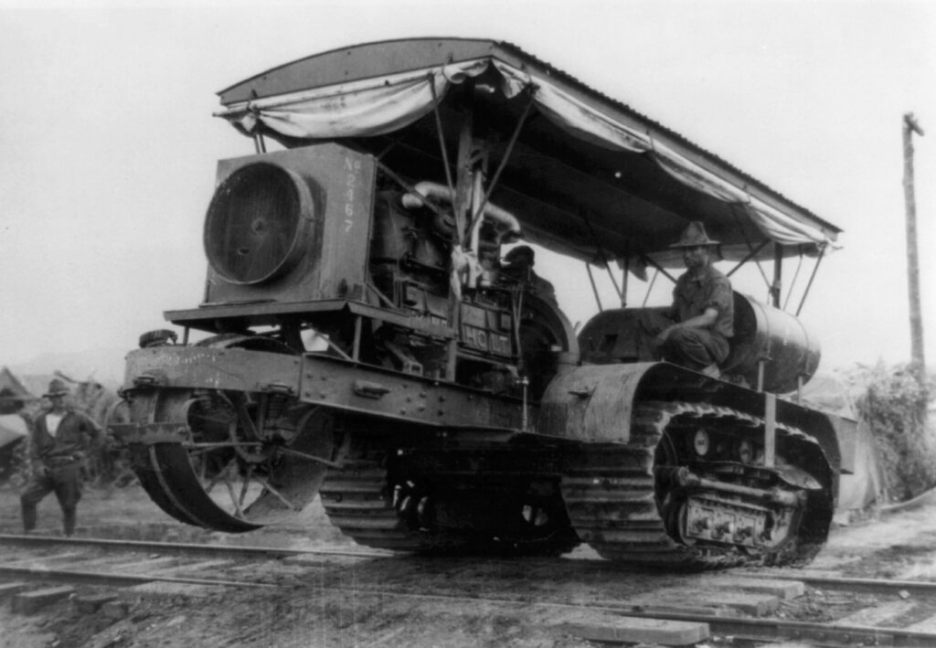 A large vehicle with tracks crossing railroad tracks, with its front wheel in the air.