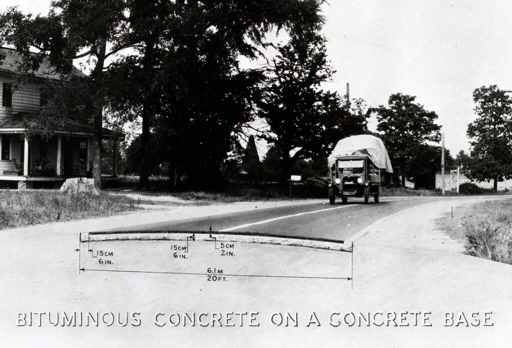 A photograph of a truck on a road with a house on the left. A cross section of the road is shown in the middle of the image. Text at the bottom of the photograph reads: “Bituminous Concrete on a Concrete Base.”