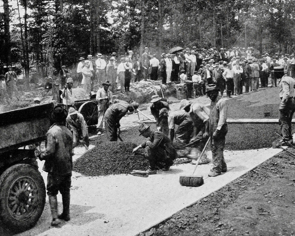 A group of men building a road while a crowd watches in the background.