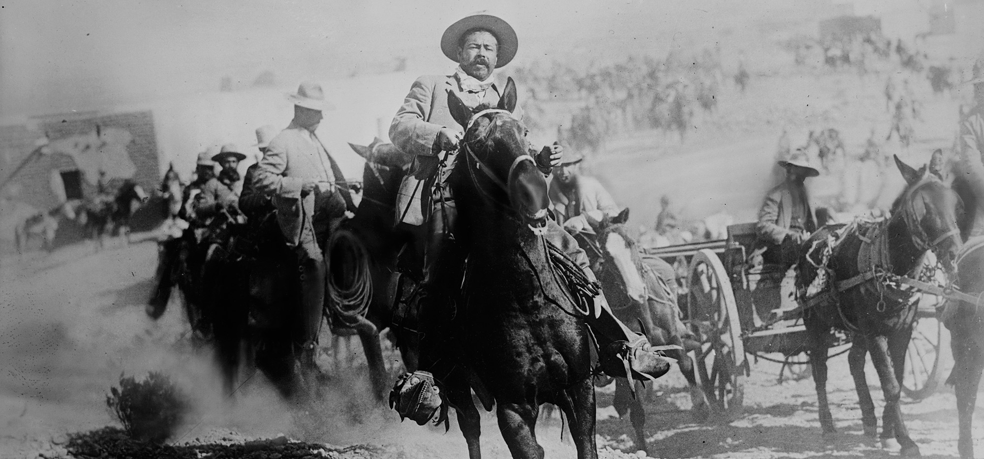 A man riding on horseback with wagons and other riders in the background.